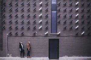 Two women standind before a wall full of mounted CCTV Cams and looking up to them in an unbelievingly way